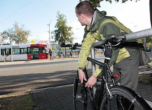 Mann schließt Fahrrad vom Fahrradbügel ab und blickt auf Straßenbahn im Hintergrund